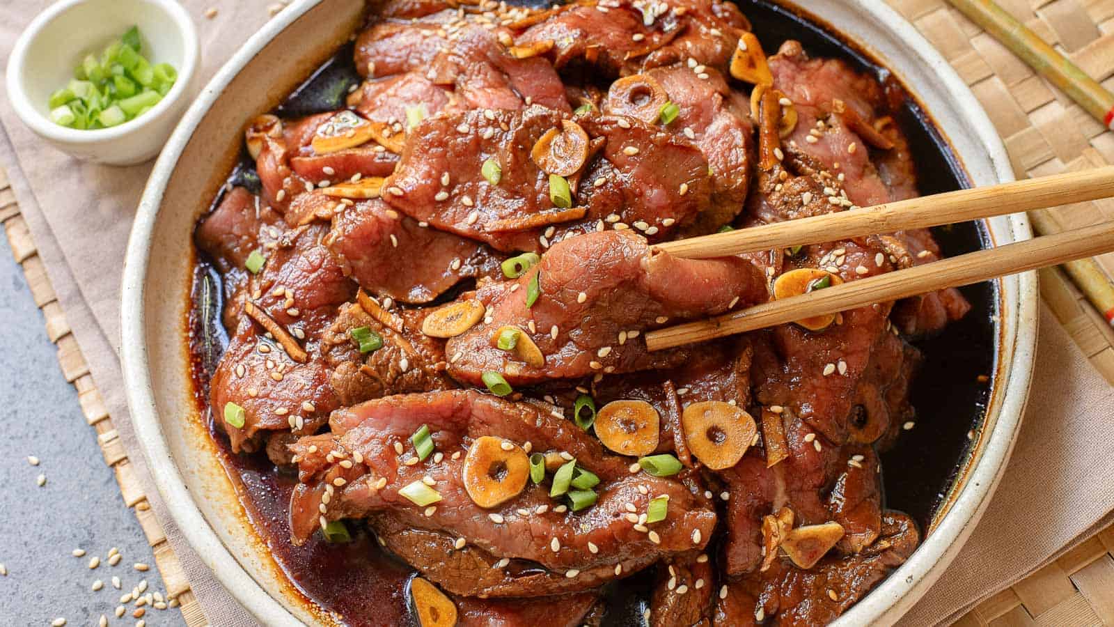 A plate of marinated beef slices topped with sesame seeds, sliced garlic, and chopped green onions, with chopsticks picking up a piece.