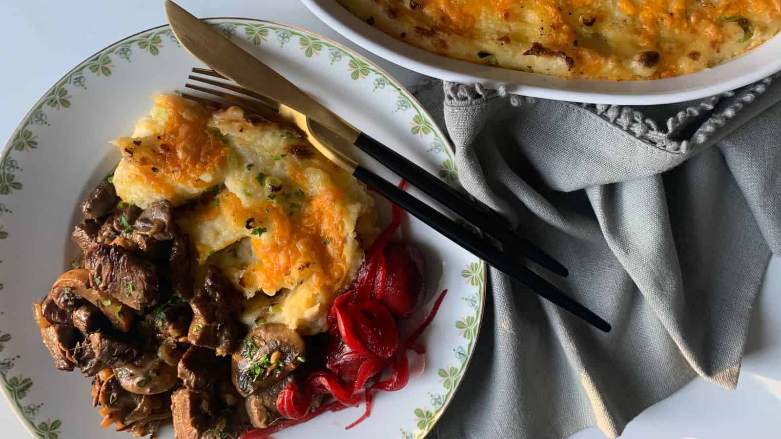 A plate with beef and mushroom stew, pickled onions, and cheesy mashed potato bake, next to a baking dish with more potato bake and a gray napkin.