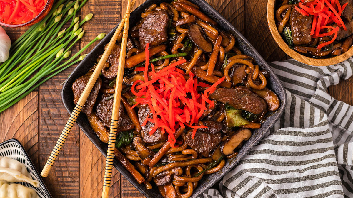A bowl of stir-fried udon noodles with beef, mushrooms, and vegetables, topped with pickled ginger and served with chopsticks on a striped cloth.