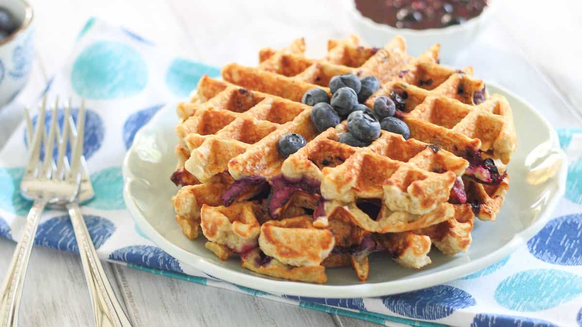 A stack of golden-brown waffles topped with fresh blueberries on a plate, with forks and a bowl of sauce in the background.