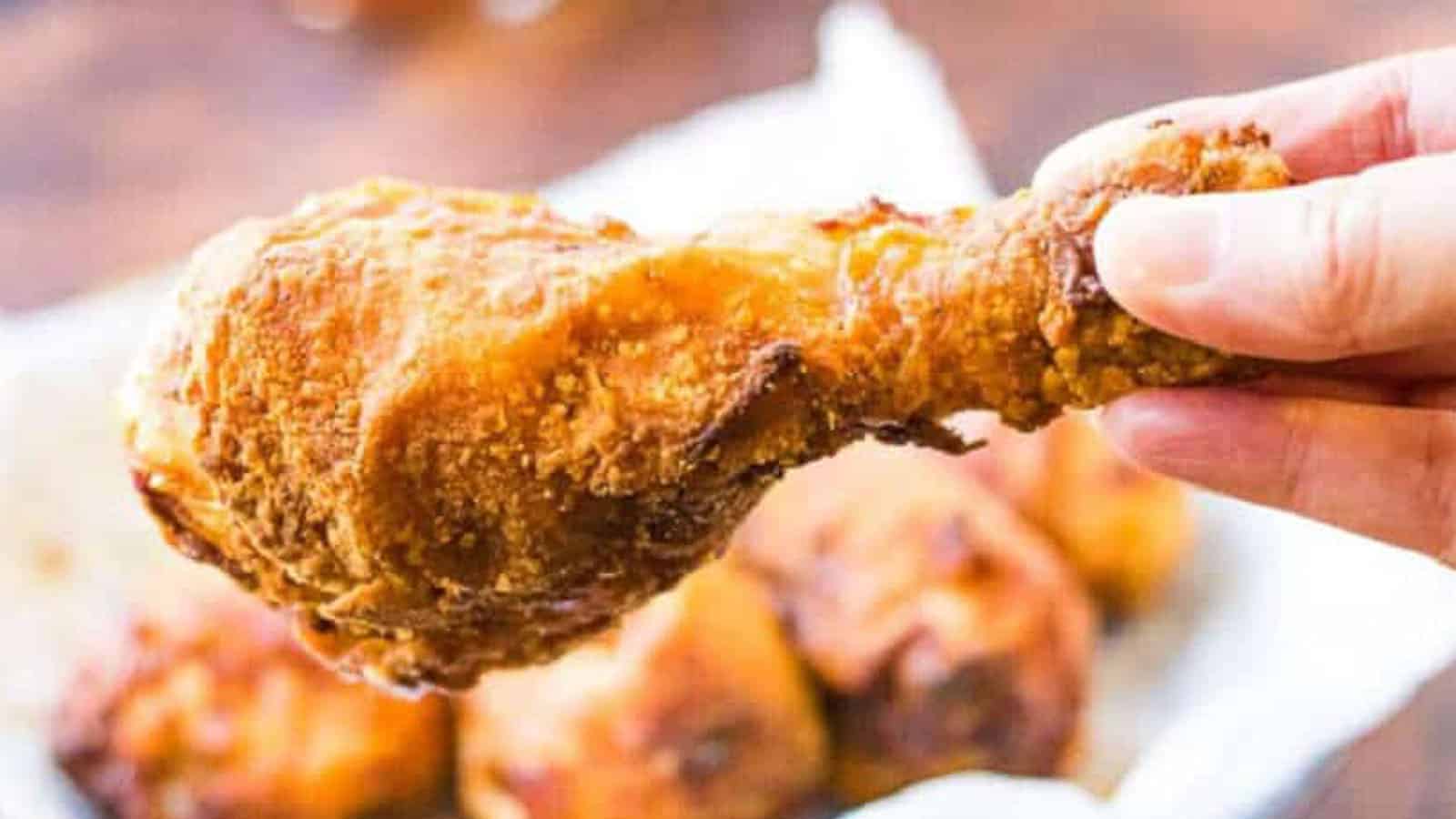 A hand holding a piece of crispy fried chicken drumstick with more pieces of fried chicken in the background.