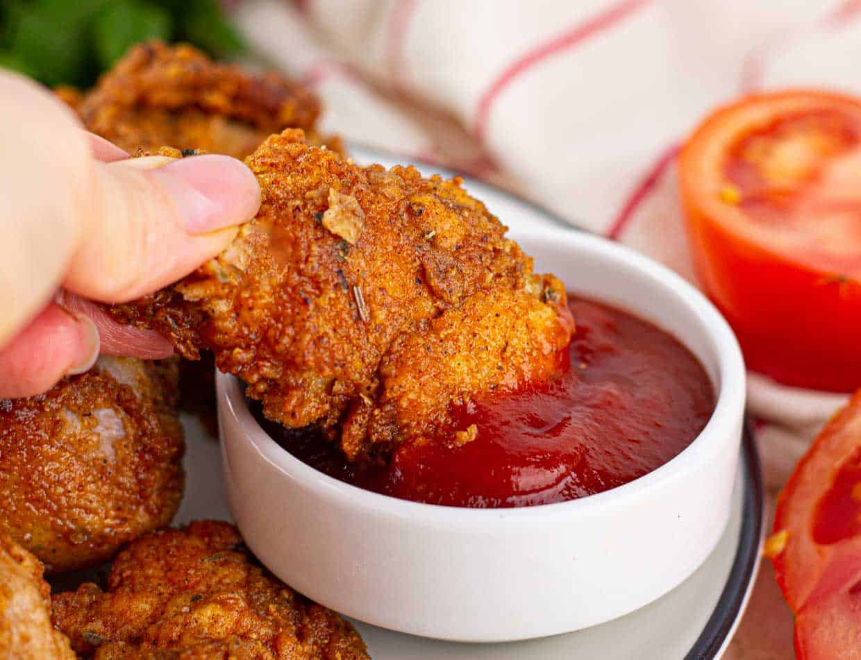 A hand dips a piece of fried chicken into a small bowl of red sauce, with more fried chicken and a sliced tomato nearby.