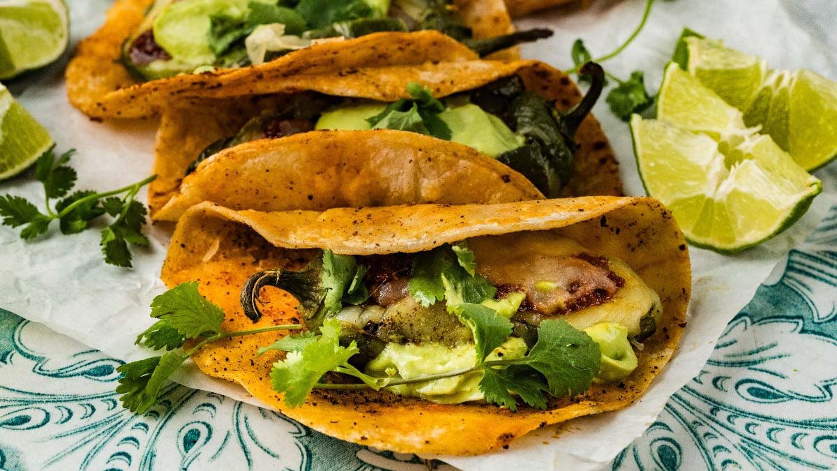 Three tacos filled with avocado, cheese, and cilantro on a patterned plate, garnished with lime wedges and fresh cilantro.