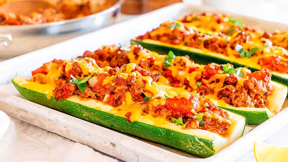 Baked zucchini boats filled with ground meat, tomatoes, and melted cheese, garnished with herbs, arranged on a white baking sheet.