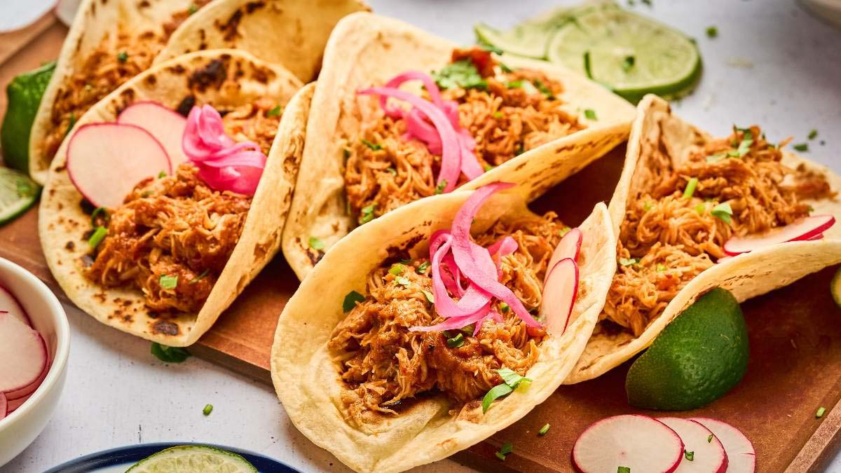 Four shredded chicken tacos in flour tortillas topped with pickled onions and radish slices, garnished with lime wedges and chopped herbs on a wooden board.