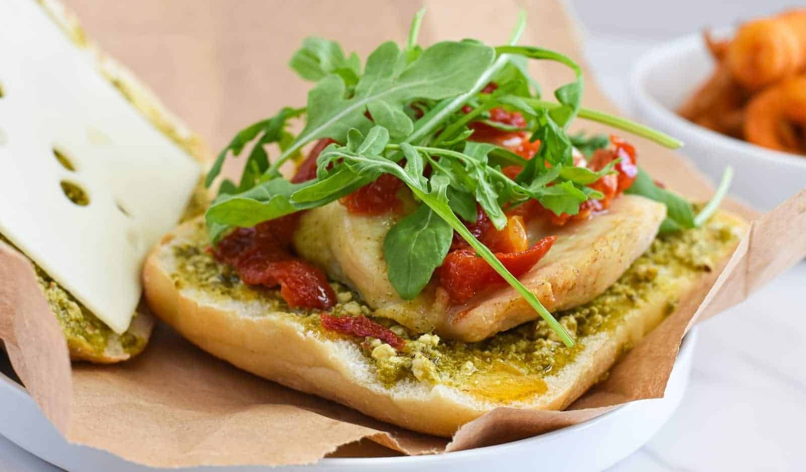 A sandwich with grilled chicken, pesto, roasted red peppers, arugula, and Swiss cheese on a hoagie roll, served on brown paper with curly fries in the background.