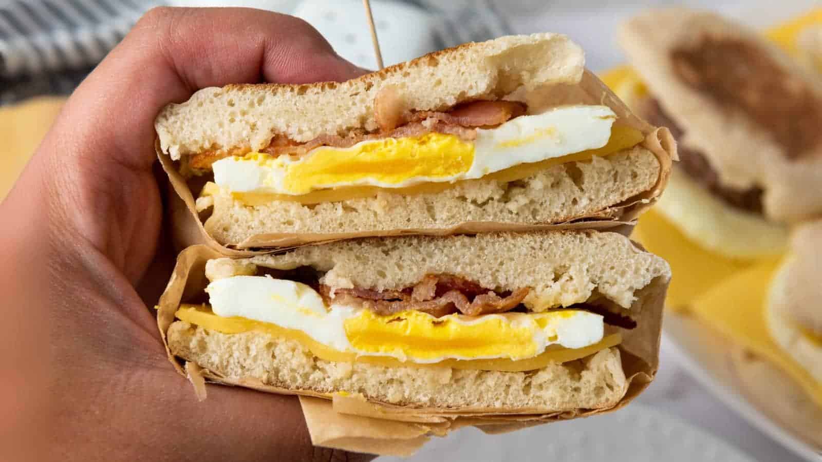 A hand holds a stacked breakfast sandwich with egg, bacon, and cheese between halves of an English muffin.