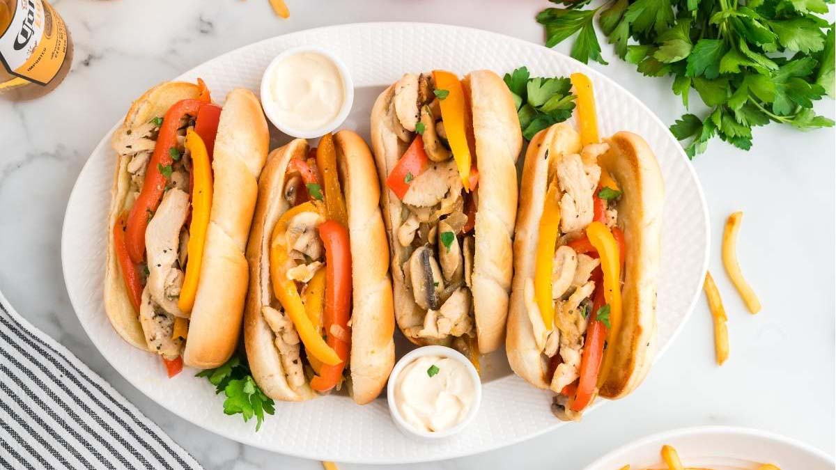 Three chicken Philly cheesesteak sandwiches with bell peppers and onions on a white platter, served with two small cups of mayonnaise.
