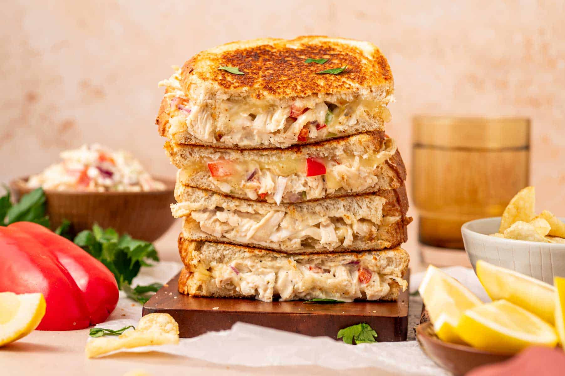A stack of three grilled sandwiches filled with a creamy mixture, surrounded by lemon wedges, potato chips, a red bell pepper, and a bowl of chopped salad.
