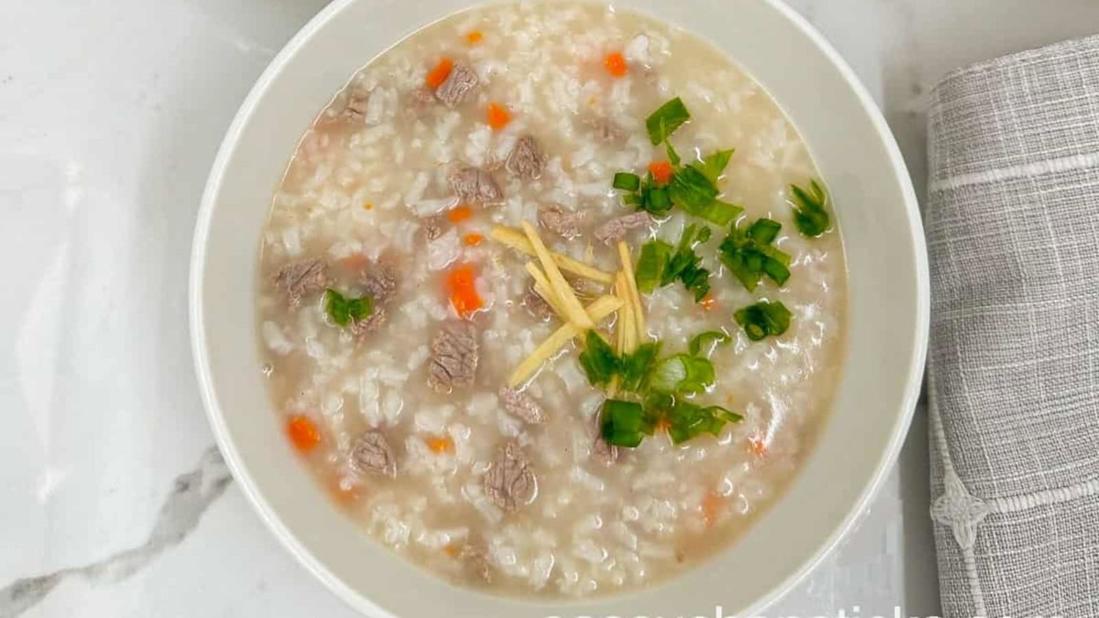 A bowl of rice porridge with diced beef, carrots, chopped green onions, and thin ginger strips, placed on a white surface beside a gray cloth.