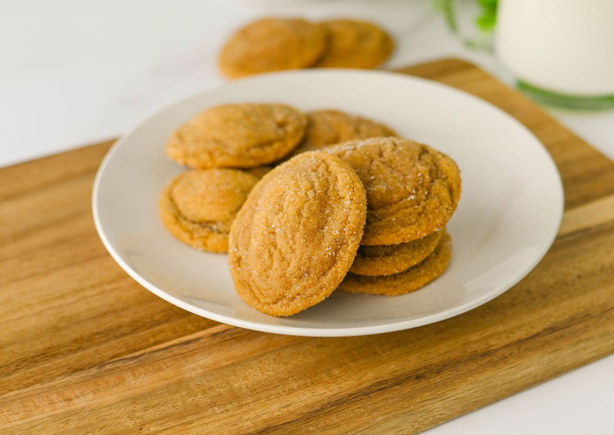 A white plate with a stack of sugar-coated cookies sits on a wooden surface, with more cookies and a glass of milk in the background.