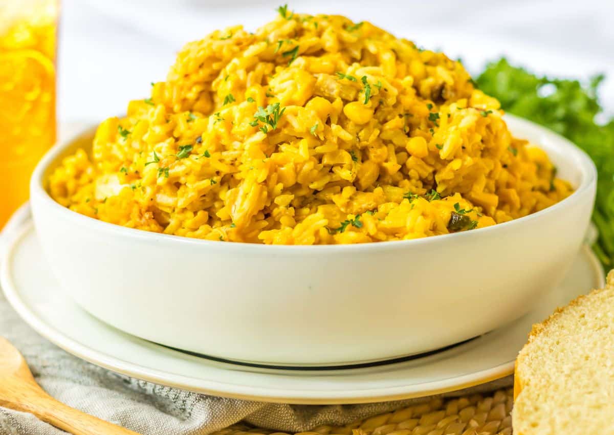 A white bowl filled with yellow rice and lentils, garnished with chopped herbs, sits on a plate next to a slice of bread and bunch of fresh greens.