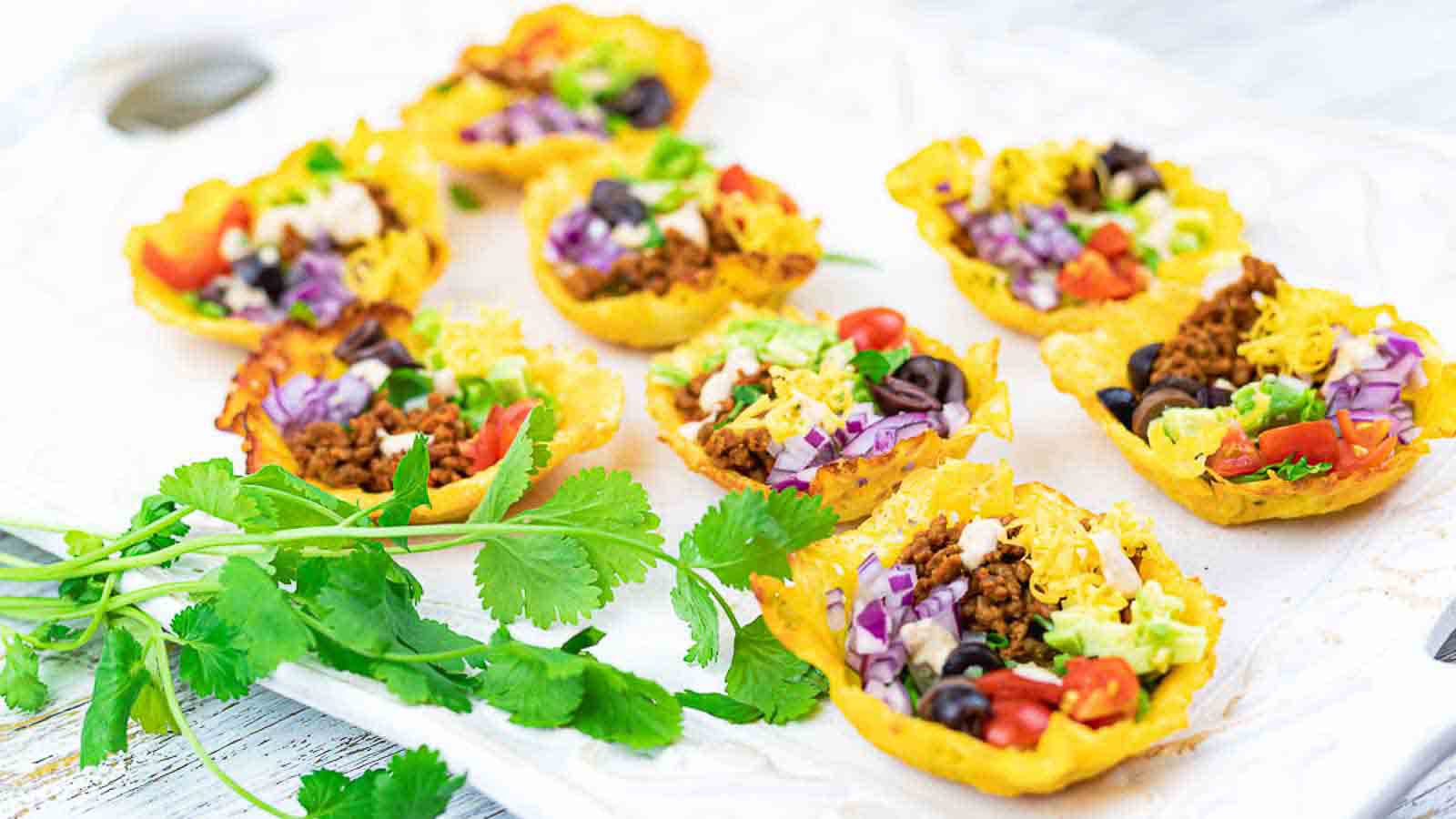 Mini taco cups filled with ground meat, lettuce, tomatoes, olives, cheese, and purple cabbage, arranged on a white tray with fresh cilantro on the side.