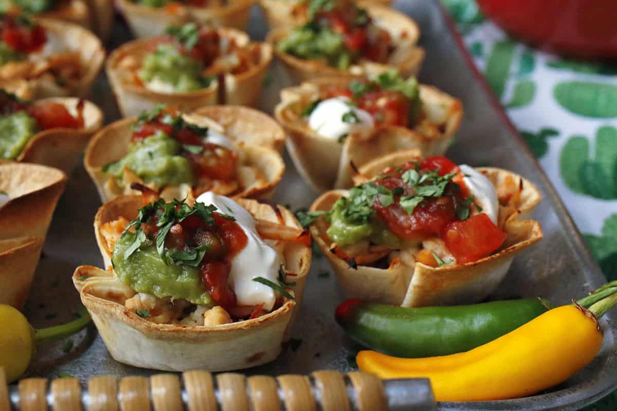 Mini tortilla cups filled with beans, cheese, salsa, guacamole, and sour cream, garnished with chopped herbs, with green and yellow chili peppers on the side.