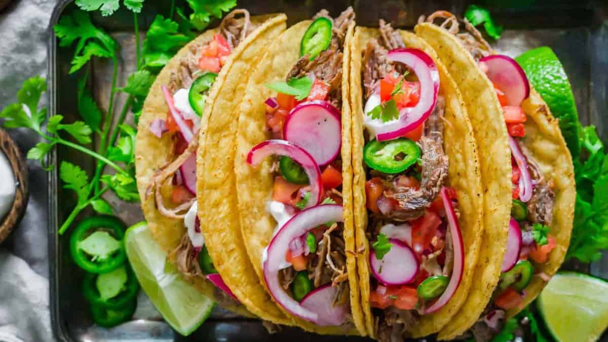 Three beef tacos with shredded meat, sliced radishes, red onions, chopped tomatoes, jalapeรฑos, and fresh cilantro, served with lime wedges on the side.