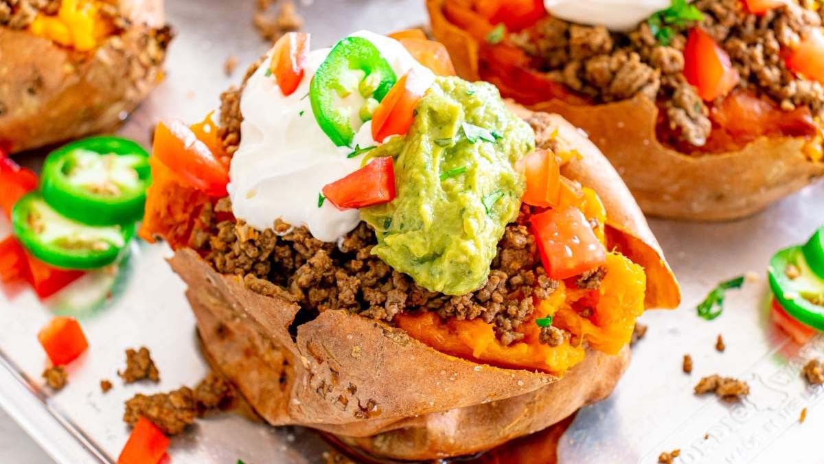 A baked sweet potato topped with ground beef, diced tomatoes, guacamole, sour cream, and sliced jalapeรฑos on a metal tray.