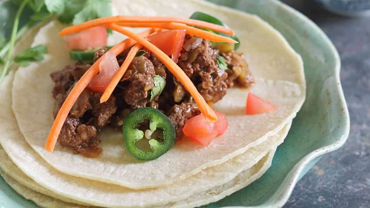 Three corn tortillas topped with seasoned ground beef, sliced carrots, jalapeรฑo, chopped tomatoes, and fresh cilantro, served on a green plate.