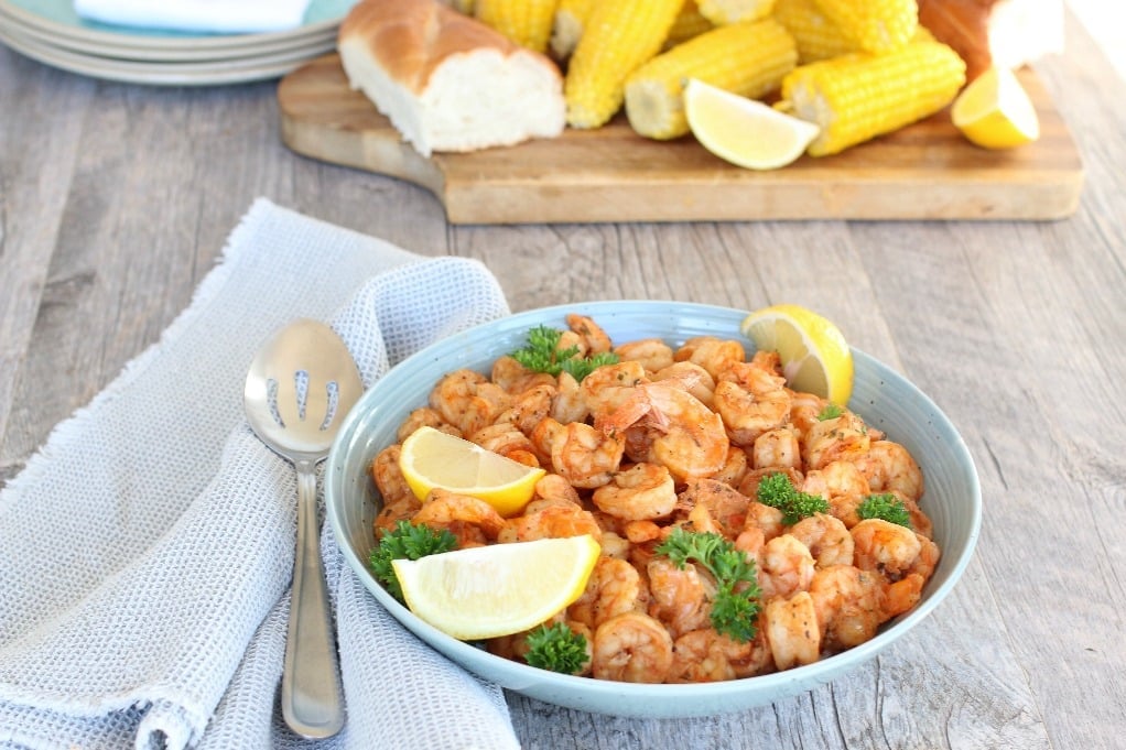 A bowl of seasoned shrimp garnished with lemon wedges and parsley, with corn on the cob and bread in the background on a wooden table.