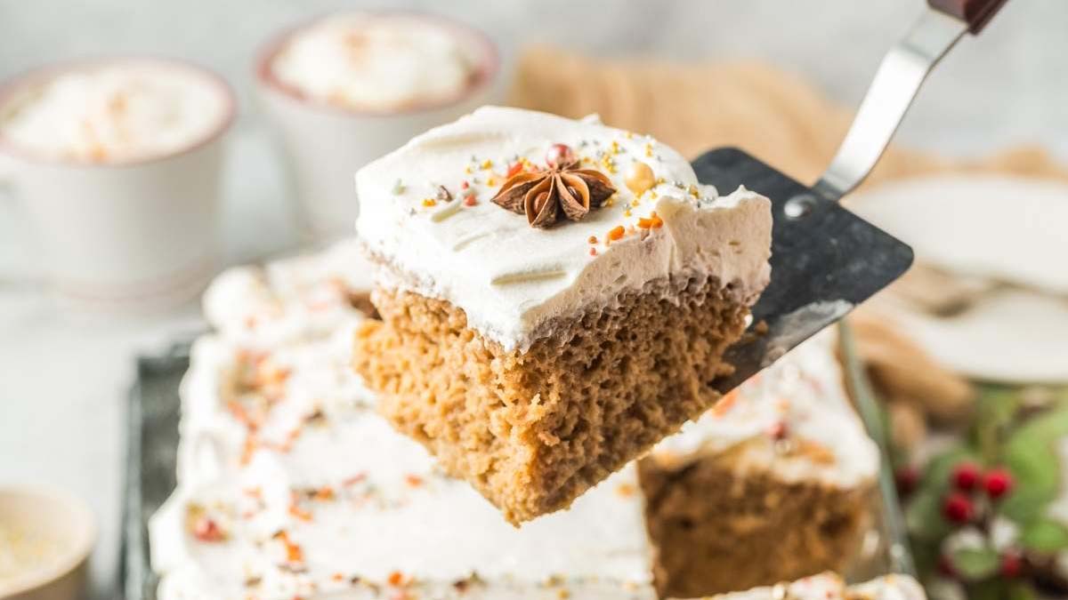 A square piece of spiced cake with white frosting and star anise garnish on a spatula, with the rest of the cake and cups in the background.