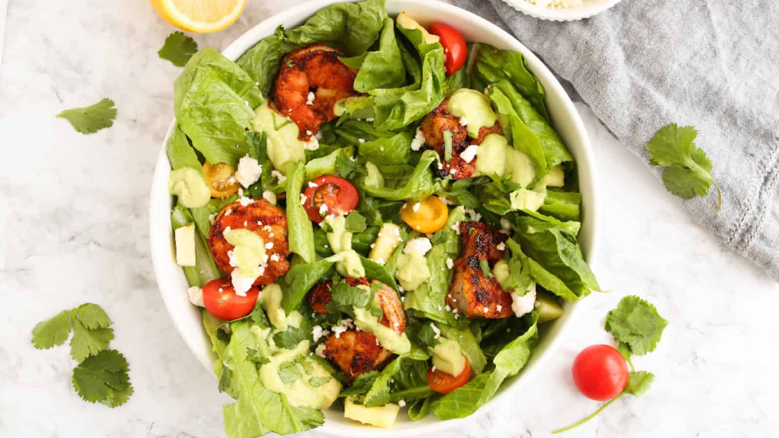 A bowl of salad with grilled shrimp, cherry tomatoes, avocado, lettuce, cheese crumbles, and green dressing, garnished with cilantro on a marble surface.