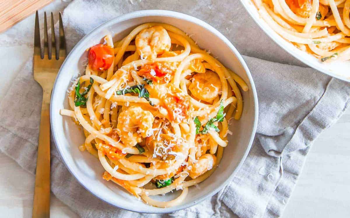 A bowl of pasta with chicken, cherry tomatoes, spinach, and grated cheese sits on a gray napkin next to a gold fork.