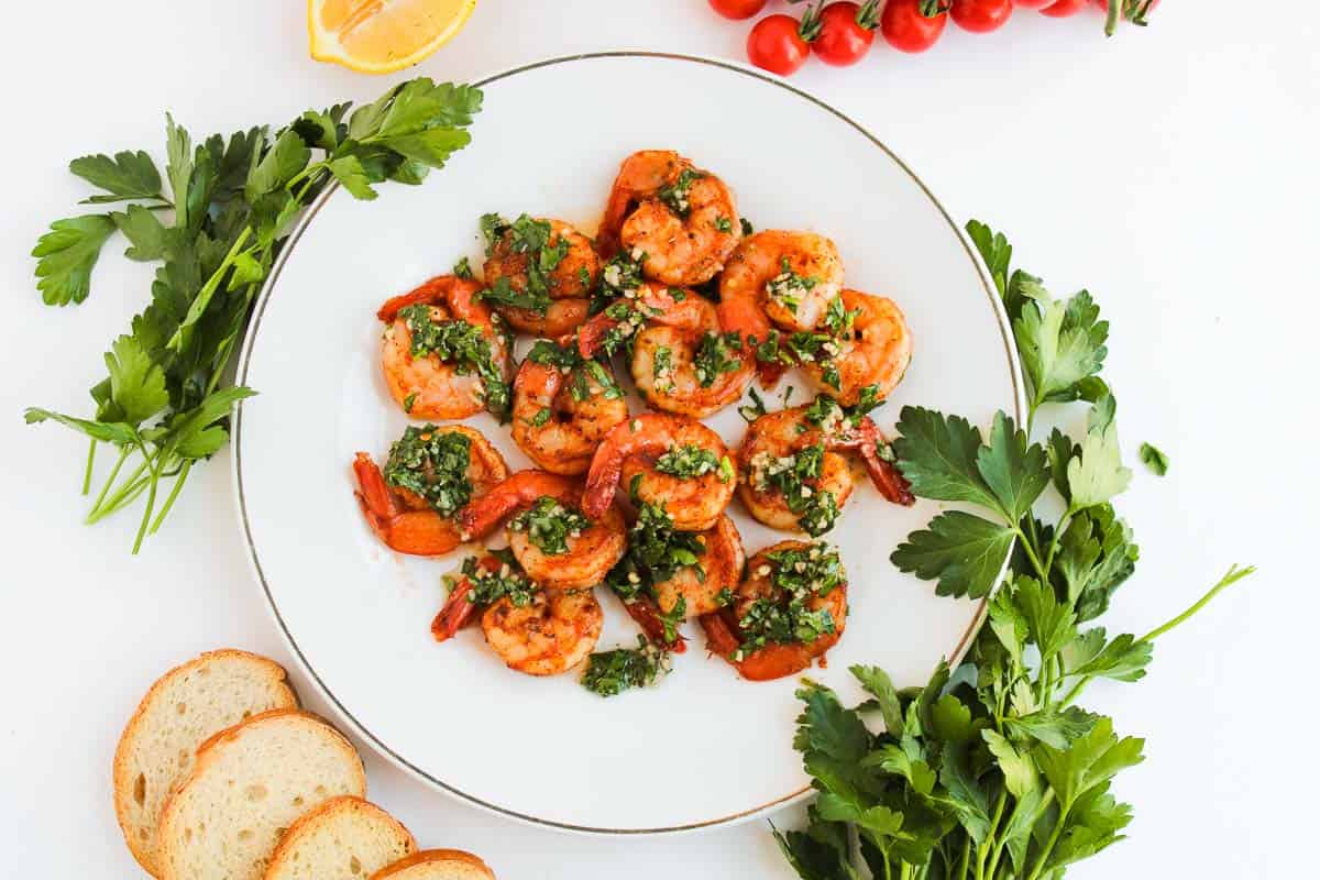 A plate of cooked shrimp garnished with green herbs, surrounded by parsley, cherry tomatoes, a lemon wedge, and slices of bread on a white surface.