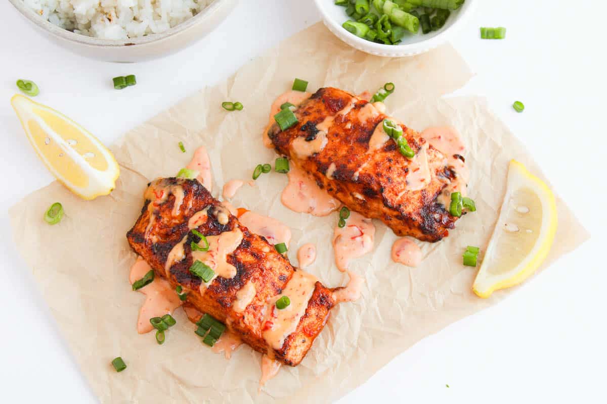 Two grilled salmon fillets topped with creamy sauce and chopped green onions, served on parchment with lemon wedges. Rice and green onions are in bowls nearby.