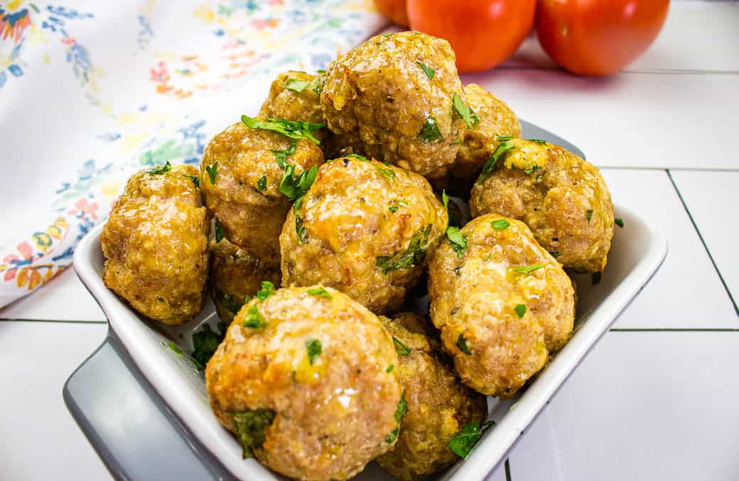 A white dish filled with homemade meatballs garnished with chopped parsley, placed on a white tiled surface with tomatoes and a floral napkin in the background.