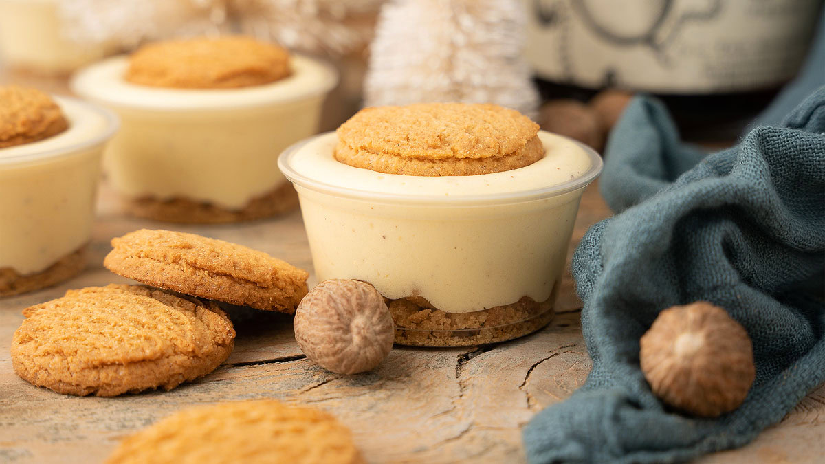 A small dessert cup filled with creamy custard, topped and layered with cookies, sits on a rustic wooden surface beside whole nutmegs and a blue cloth.