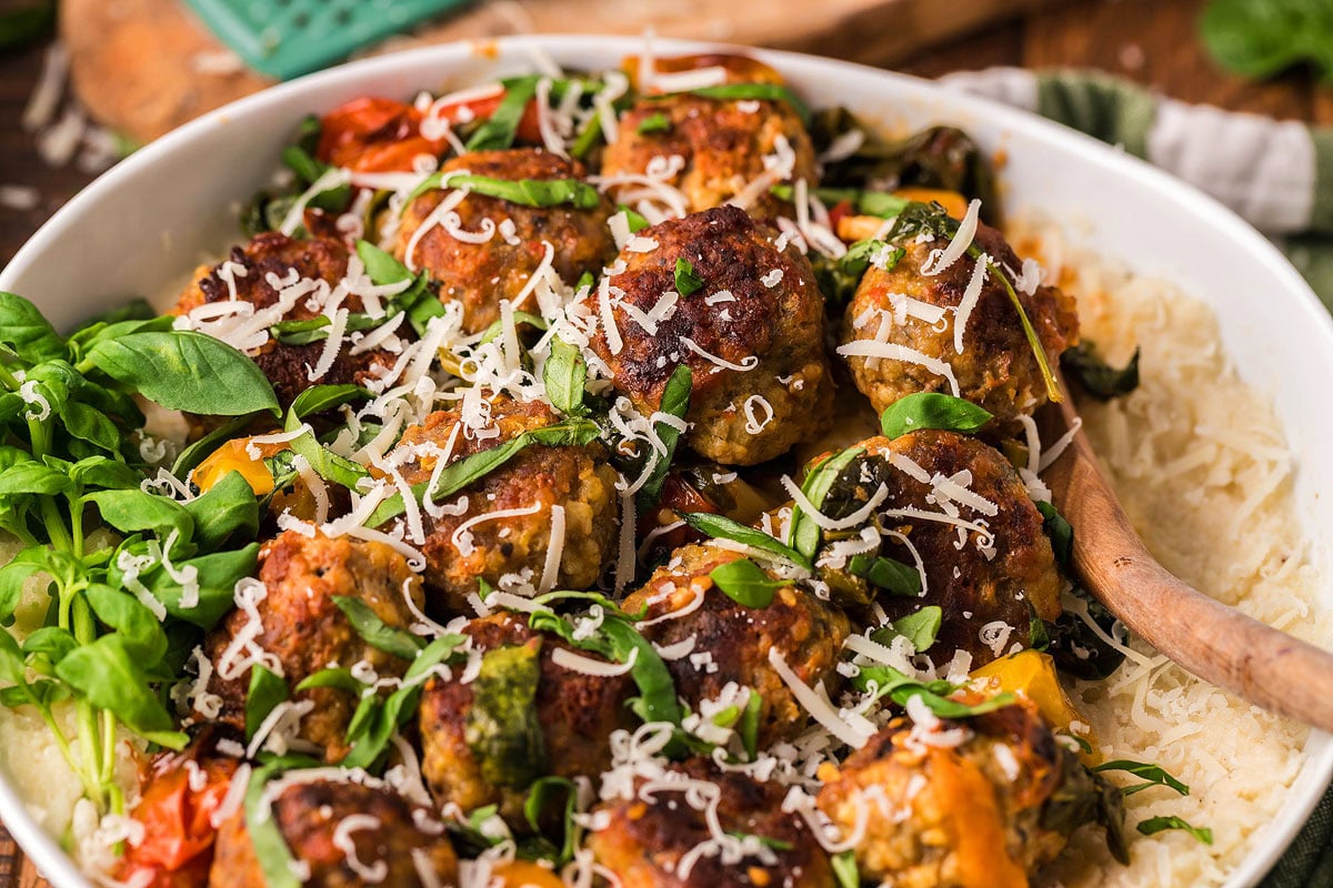 A bowl of meatballs topped with grated cheese and fresh basil, served on a bed of rice or shredded vegetables with a wooden spoon.