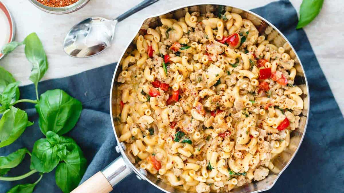 A pan filled with cooked macaroni mixed with ground meat, spinach, cherry tomatoes, and herbs sits on a dark cloth next to fresh basil leaves.