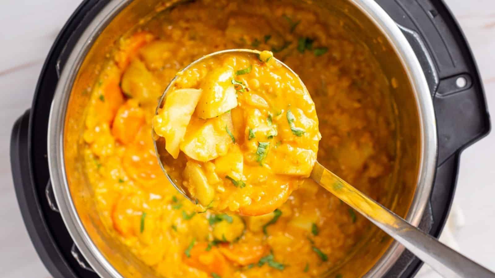 A ladle lifts a serving of thick, yellow vegetable curry with visible herbs and potato chunks from a pot.