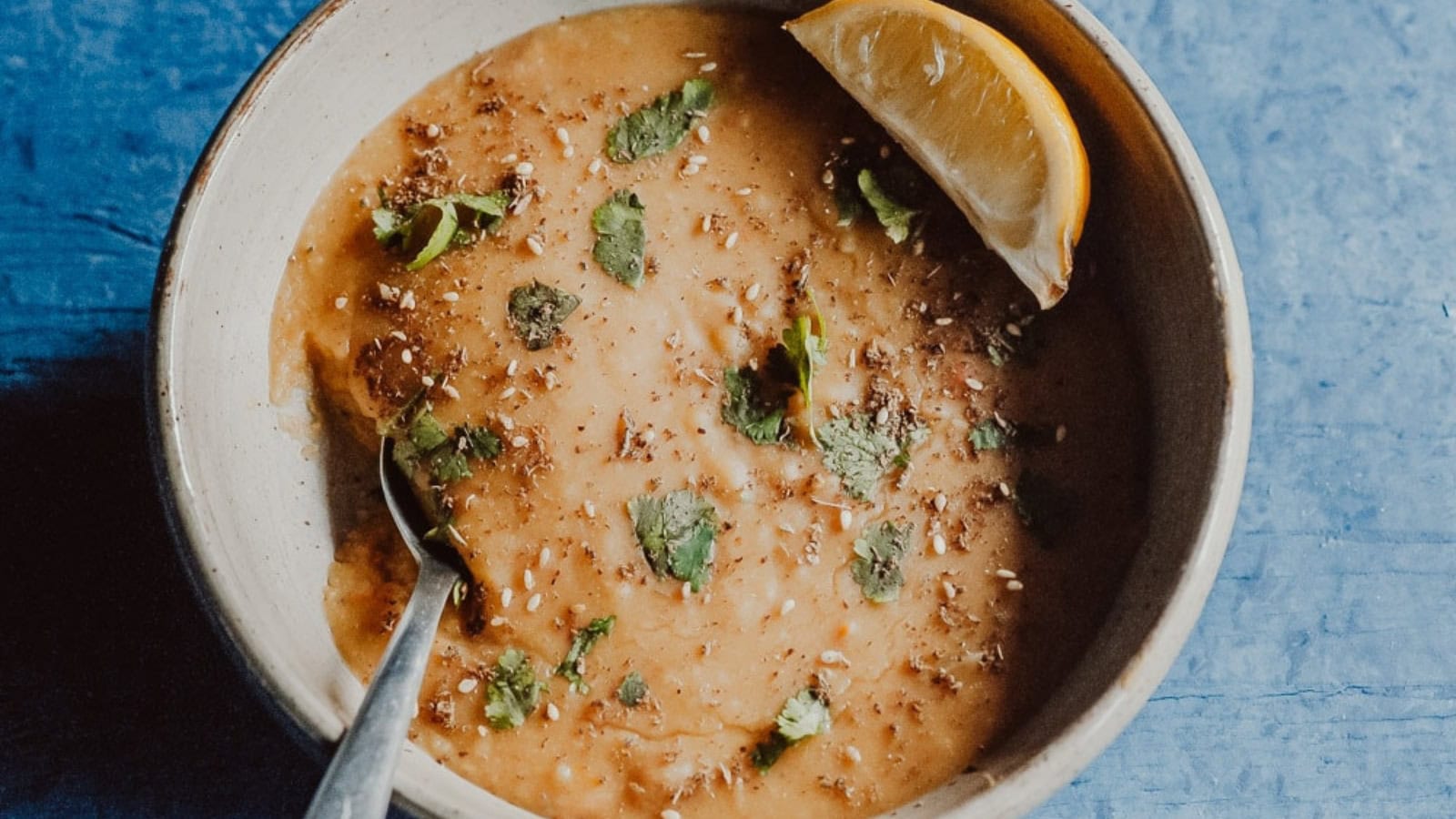 A bowl of thick soup garnished with herbs and spices, served with a lemon wedge on the rim, and a spoon resting inside.