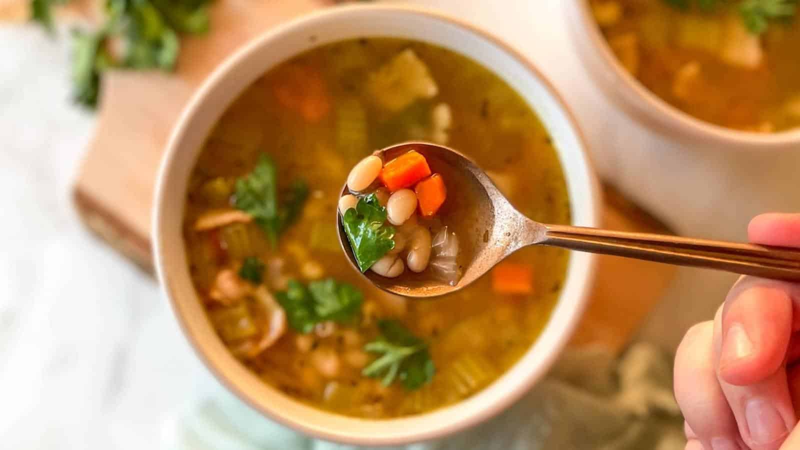 A hand holds a spoonful of bean and vegetable soup above a bowl filled with the same soup, containing beans, carrots, celery, and herbs.