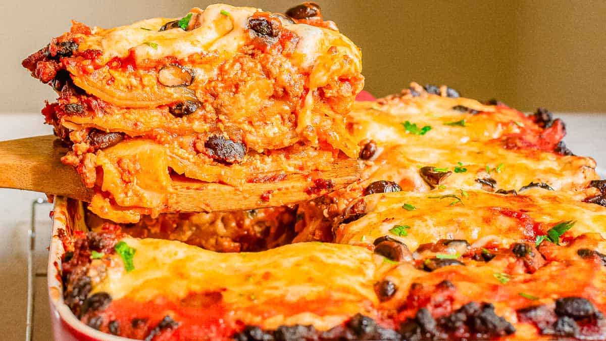 A close-up of a baked lasagna being lifted from a dish, showing layers of pasta, cheese, tomato sauce, and black olives.