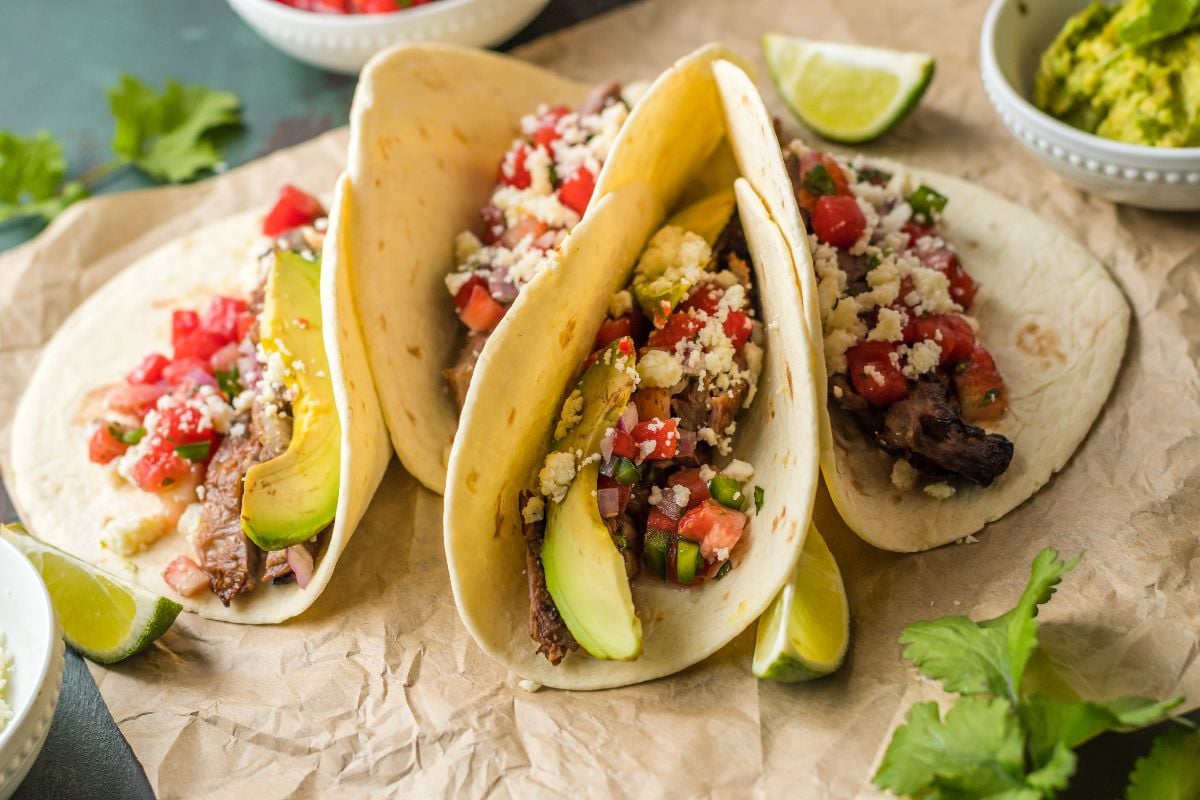 Three soft tacos filled with grilled meat, sliced avocado, diced tomatoes, cheese, and herbs, served on parchment paper with a lime wedge and cilantro garnish.