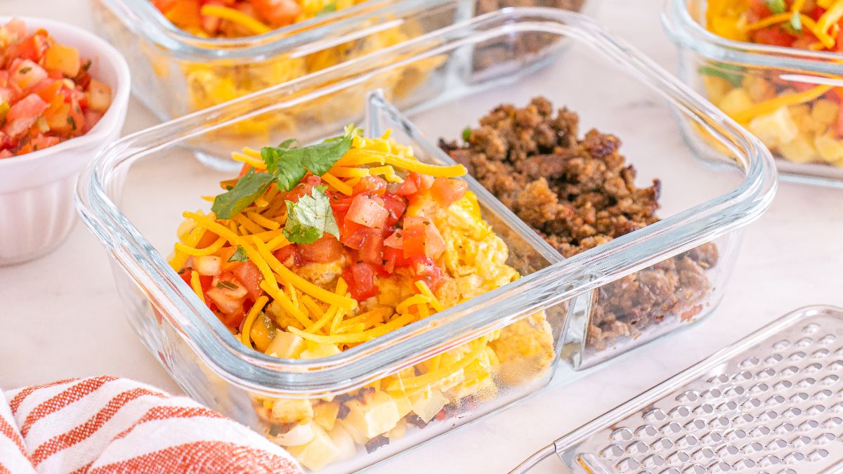 A glass meal prep container holds scrambled eggs topped with shredded cheese and diced tomatoes, with cooked ground beef in a separate section. Other similar containers are visible in the background.
