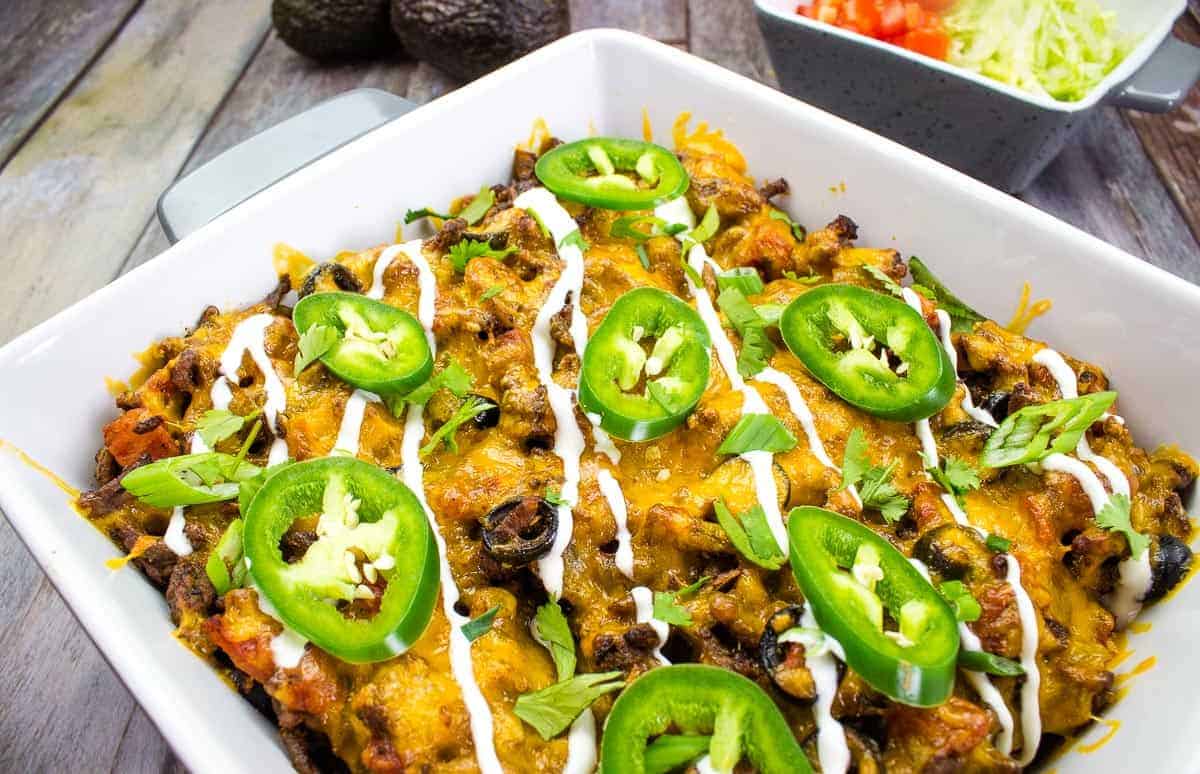 A baked casserole topped with sliced jalapeรฑos, chopped green onions, melted cheese, and drizzled with sour cream. A bowl of shredded lettuce and diced tomatoes is in the background.