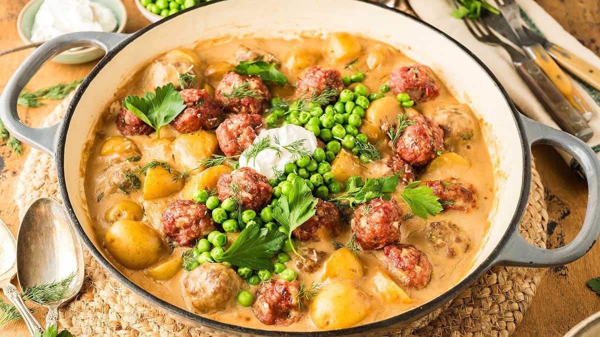 A cast iron skillet filled with meatballs, baby potatoes, peas, and creamy sauce, garnished with fresh herbs and a dollop of sour cream.