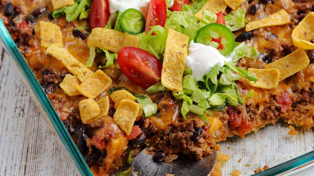 A glass baking dish filled with taco casserole topped with corn chips, shredded lettuce, sliced tomatoes, jalapeรฑos, and sour cream. A serving has been scooped out with a spoon.