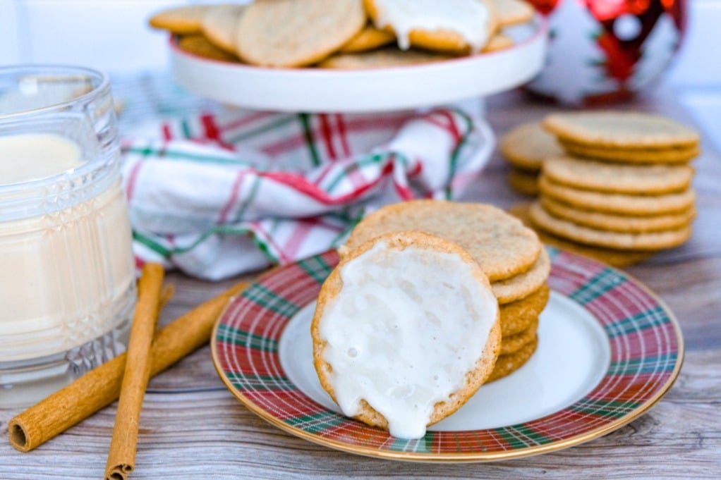 A plate of cookies, one with white icing, sits next to a glass of milk, cinnamon sticks, and more cookies on a tray; a plaid cloth is in the background.