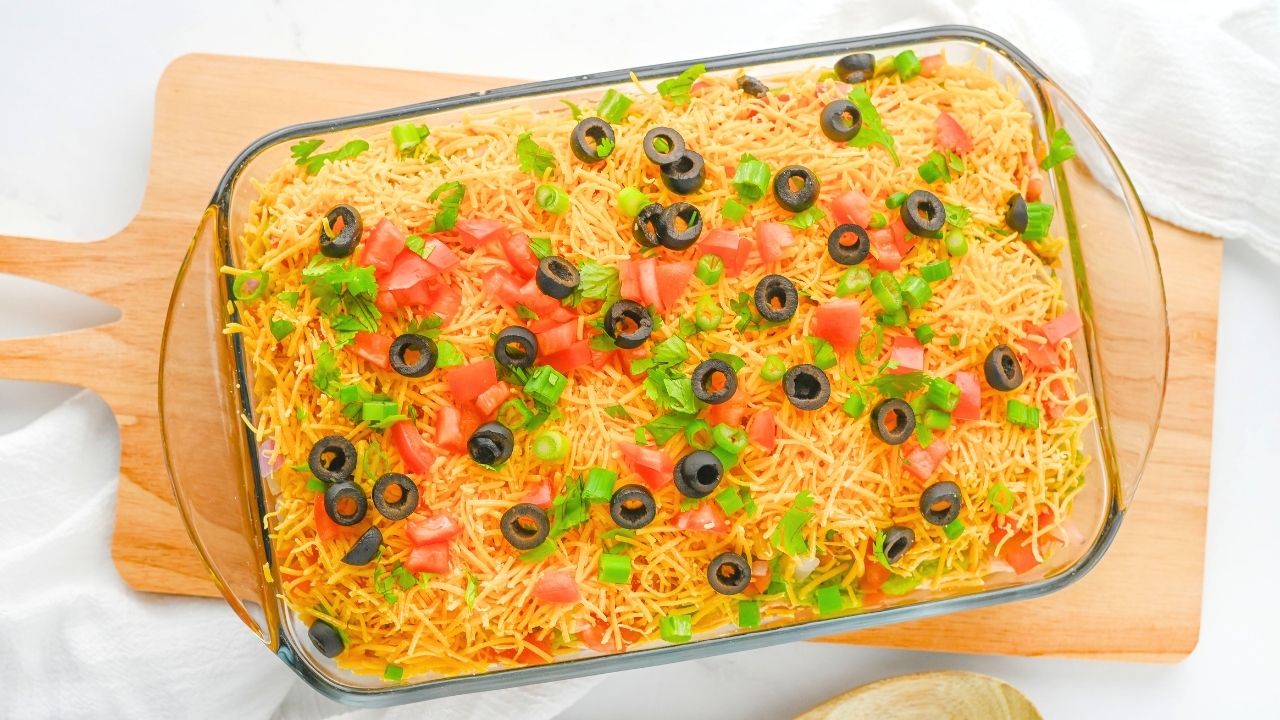 A glass baking dish filled with layered dip, topped with shredded cheese, chopped tomatoes, sliced black olives, and green onions on a wooden board.