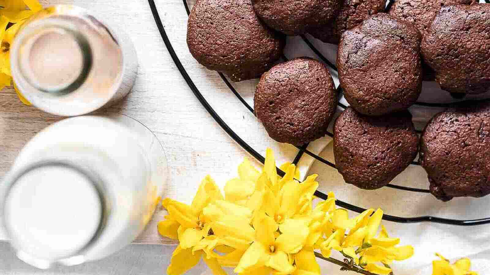 A cooling rack with chocolate cookies next to yellow flowers and two glass bottles of milk on a light surface.