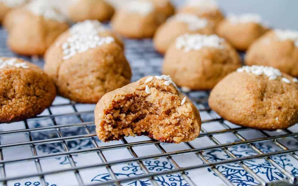 Close-up of several brown cookies with white sugar sprinkles on a cooling rack, one with a bite taken out showing the soft interior.