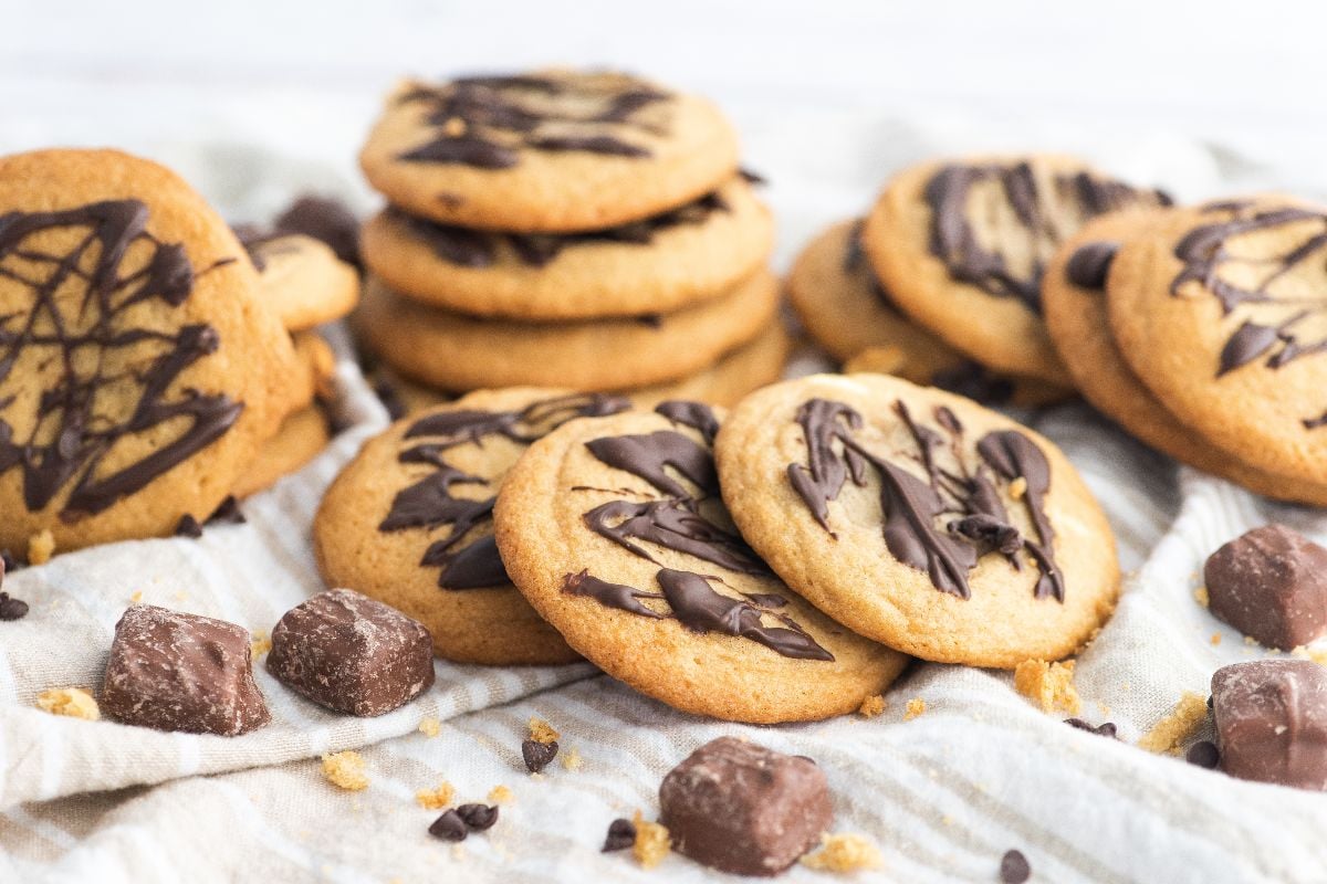 A group of cookies drizzled with chocolate are arranged on a cloth alongside chocolate chunks and crumbs.