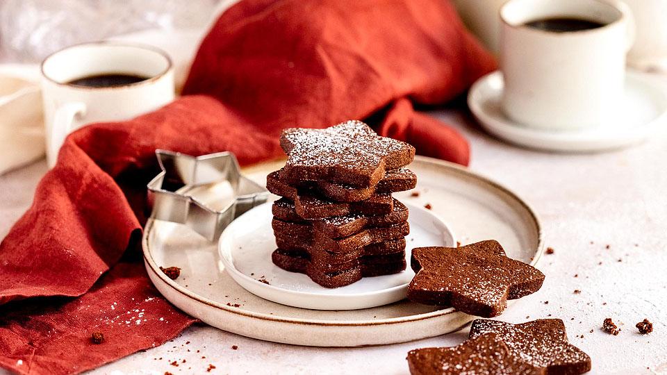 A stack of star-shaped chocolate cookies dusted with powdered sugar on a plate, with a cookie cutter, cups of coffee, and a red napkin in the background.