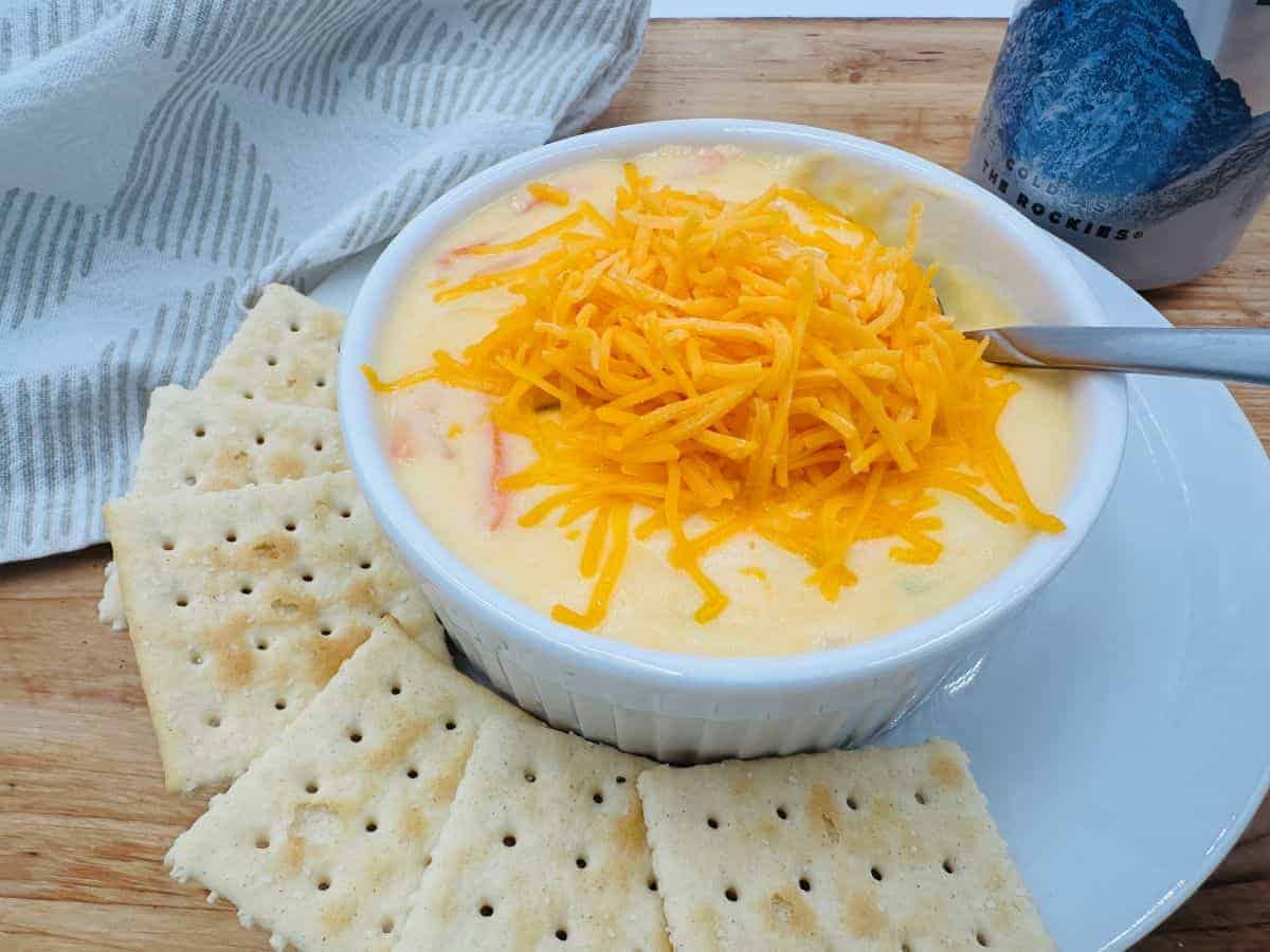 A bowl of cheesy soup topped with shredded cheddar, surrounded by saltine crackers on a white plate, with a spoon and a striped towel nearby.