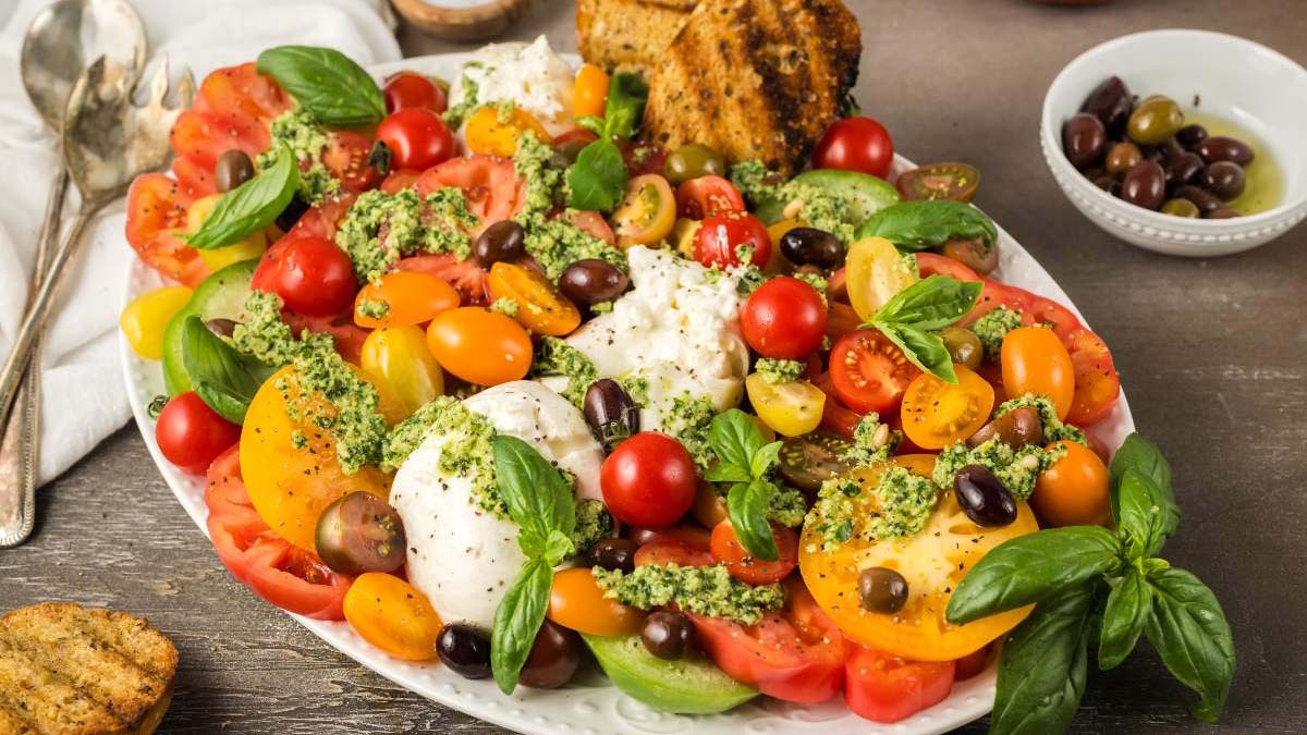 A platter of colorful heirloom tomatoes, mozzarella balls, olives, fresh basil, pesto, and toasted bread slices on a wooden table.