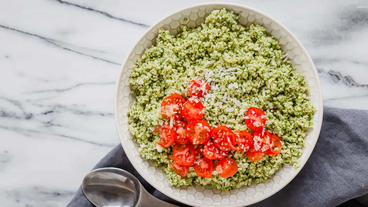 A bowl of green-colored couscous topped with chopped cherry tomatoes and grated cheese, placed on a marble surface next to a spoon.