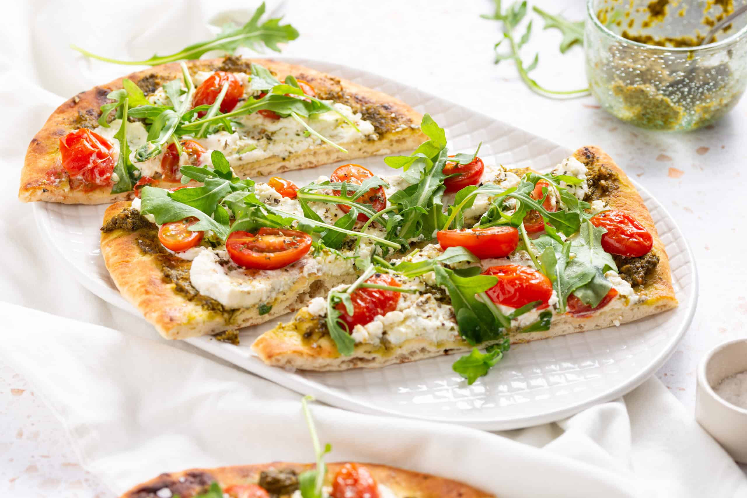A rectangular plate with three slices of pizza topped with arugula, cherry tomatoes, ricotta cheese, and pesto sauce.